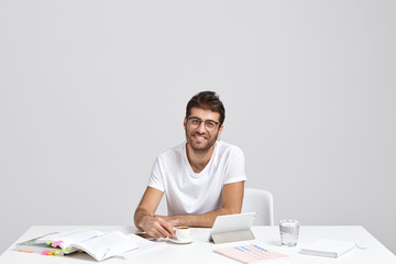 Prosperous smiling male entrepreneur in white t shirt, drinks coffee, wears transparent glasses, smiles gladfully, sits at desktop with tablet, works on new startup based on innovative forecasting