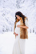 © gorynvd - Beautiful pregnant woman in a long light dress and coat gently holds her stomach with her hands in a snowy winter park on background.