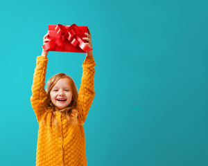 Portrait of a little girl in a warm mustard knitted sweater on a cyan background. A child holds a red box with a gift above his head. The concept of celebration, giving and receiving a gift.