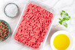 © laplateresca - A photo of minced meat in a plastic tray, shot from the top on a white marble kitchen surface with salt, pepper, olive oil, and parsley leaves and copy space
