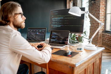 young male programmer writing a program code sitting at the workplace with three monitors in the off