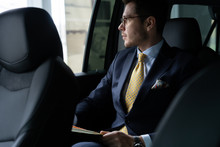Young Businessman Sitting On Back Seat Of The Car, While His Chauffeur Is Driving Automobile.