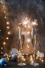 Two Glasses Of Sparkling Wine With Sparklers. Dark Background With Yellow Light Bokeh. Christmas Tree Toys On The Table.