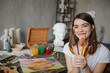 © Iryna&Maya - Adorable young artist girl holding brushes and smiling. Creative workshop room at the background