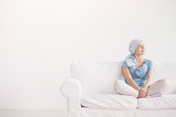 Wall Mural - Happy teenage girl suffering from breast cancer, wearing blue headscarf and siting cross leg on the couch at home before treatment, copy space on empty wall