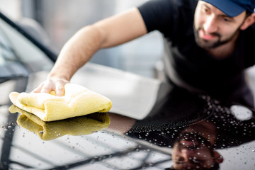 Professional washer in t-shirt and cap wiping a car hood with yellow microfiber at the open air car wash