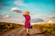 © Masson - Redhead girl in red dress with hat and suitcase walking down on rural road near wheat field and cloudscape on background.