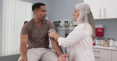 Wall Mural - Senior female doctor checking young black males blood pressure with monitor and stethoscope. Mature caucasian doctor checking blood pressure of young african-american patient