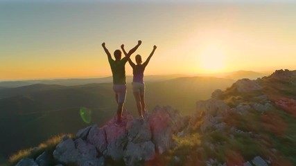 Wall Mural - DRONE, LENS FLARE: Flying over sporty couple outstretching arms after a successful hike in the scenic mountains in Slovenia. Cheerful young tourists outstretching arms on a golden summer evening.