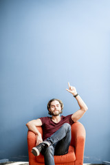 Wall Mural - Portrait of a young caucasian bearded man with long hair showing with hands on the colorful background sitting on the chair. Image with copy space