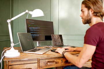 Wall Mural - young programmer writing a program code sitting at the workplace with three monitors in the office o