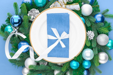 elevated view of napkin wrapped by festive ribbon on plate surrounded by evergreen tree branches with christmas balls isolated on blue