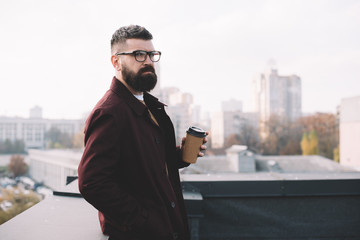 focused stylish adult man in glasses holding coffee to go on rooftop