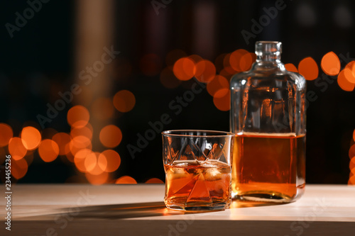 Cuadros en Lienzo Glass and bottle of whiskey on wooden table against blurred lights