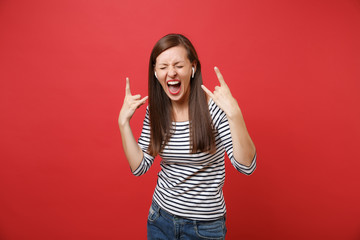 Screaming young woman with wireless earphones depicting heavy metal rock horns sign, listening music isolated on red wall background. People sincere emotions, lifestyle concept. Mock up copy space.