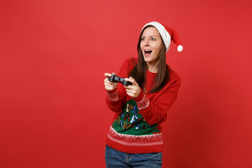 Wall Mural - Amazed young Santa girl in Christmas hat keeping mouth wide open playing video game with joystick isolated on red background. Happy New Year 2019 celebration holiday party concept. Mock up copy space.