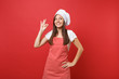 Housewife female chef cook or baker in striped apron white t-shirt, toque chefs hat isolated on red wall background. Smiling calm cute woman making okay taste delight sign. Mock up copy space concept.