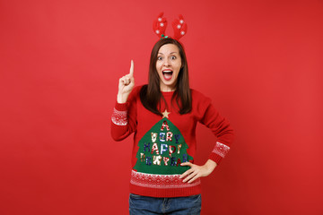 Surprised Santa girl in fun decorative deer horns holding index finger up with great new idea keeping mouth wide open isolated on red background. Happy New Year 2019 celebration holiday party concept.