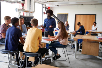 Group Of High School Students Sitting At Work Benches Listening To Teacher In Design And Technology Lesson