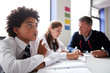 © Monkey Business - Concentrating Male High School Student Wearing Uniform Working At Table With Teacher Talking To Pupils In Background
