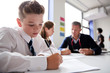 © Monkey Business - Male High School Student Wearing Uniform Working At Table With Teacher Talking To Pupils In Background