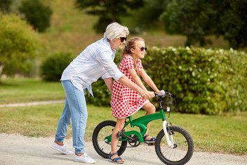 family, leisure and people concept - happy grandmother teaching granddaughter to ride bicycle at summer park