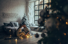 Young Couple Sitting Near Bed And Hugging On Background Of Christmas Tree, Glowing Lightbulbs And Window.