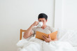 © Asada - Asian Man drinking a cup of coffee or breakfast In bed while reading the book in the morning.