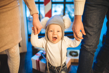 Wall Mural - Funny little baby boy 1 year old learning walk home in winter in a decorated New Year house. Young family dad and mom hold by the hands of his son in the loft interior wooden floor near the window