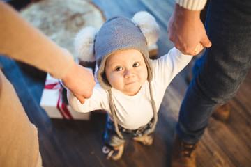 Wall Mural - Funny little baby boy 1 year old learning walk home in winter in a decorated New Year house. Young family dad and mom hold by the hands of his son in the loft interior wooden floor near the window