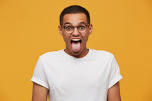 Portrait Of Attractive Young Man In Glasses, Jokes, Stuck Out His Tongue, Grimaces Looks Funny, Wears White Casual T-shirt, On A Yellow Background