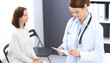 © rogerphoto - Doctor woman at work. Portrait of female physician filling up medical form while standing near reception desk at clinic or emergency hospital. Patient woman sitting at the background. Medicine concept