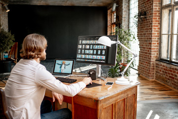 Young male photographer working with woman's portraits sitting at the working place with two computers in the studio