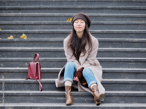 Fotografía Portrait of beautiful young woman thinking sitting on wooden steps in a park outdoors background late autumn, smiling with eyes closed, with red bag beside her