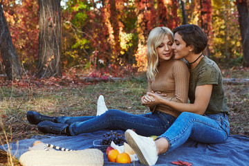 A beautiful happy couple of lesbian ladies having a romantic picnic in the park. The blonde and brunette young women sitting on the blanket, holding hands, hugging. Autumn trees in the background.