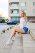 © Adriana Nikolova - Attractive blonde woman in stylish clothes sitting on stairs at street with blurred building