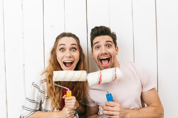 Image of funny couple man and woman holding paint rollers and standing over white wall, while painting and making renovation