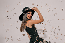 Cheerful Brunette Woman Holding Her Hat And Jumping Over Isolated Background. Carefree Girl Enjoying Confetti Around And Have A Good Mood At New Year Party.