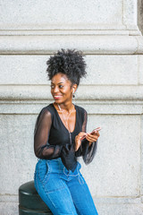 Wall Mural - Young beautiful African American Woman with afro hairstyle wearing mesh sheer long sleeve shirt blouse, sitting on street in New York, listening music with earphone and cell phone, smiling..