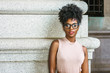 © Alexander Image - Portrait of Young African American Woman in New York. Young black female college student with afro hairstyle wearing sleeveless light color top, eye glasses, standing by column on street on campus..