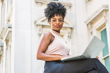 Wall Mural - Way to Success. Young African American woman with afro hairstyle wearing sleeveless light color top, sitting by vintage office building in New York, working on laptop computer, looking, thinking..