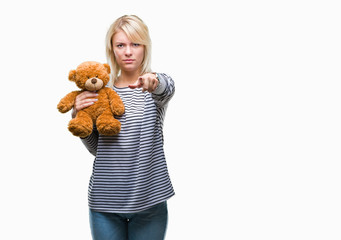 Young beautiful blonde woman holding teddy bear plush over isolated background pointing with finger to the camera and to you, hand sign, positive and confident gesture from the front