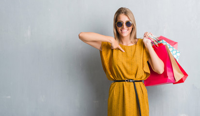 Wall Mural - Beautiful young woman over grunge grey wall holding shopping bags on sales with surprise face pointing finger to himself