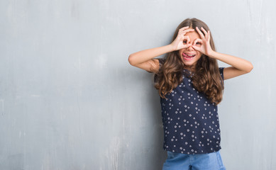 Wall Mural - Young hispanic kid over grunge grey wall doing ok gesture like binoculars sticking tongue out, eyes looking through fingers. Crazy expression.