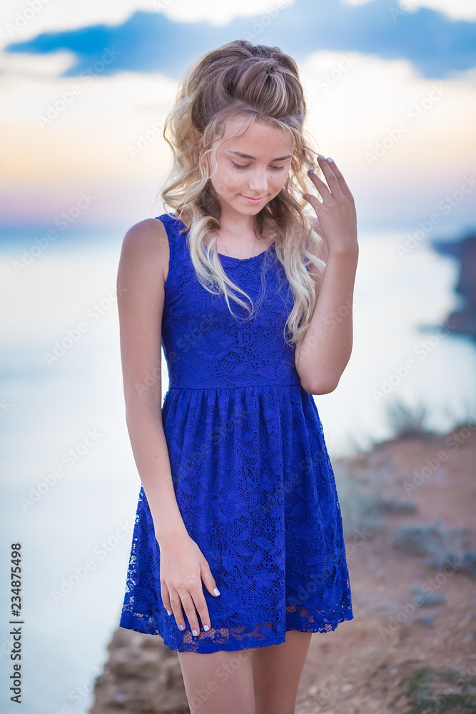 Cute young lady with curly blond hair posing on beach wearing tender blue  dress on sea side background and blue heaven sky. Adore scene of pretty  emancipated girl with tender skin. Stock