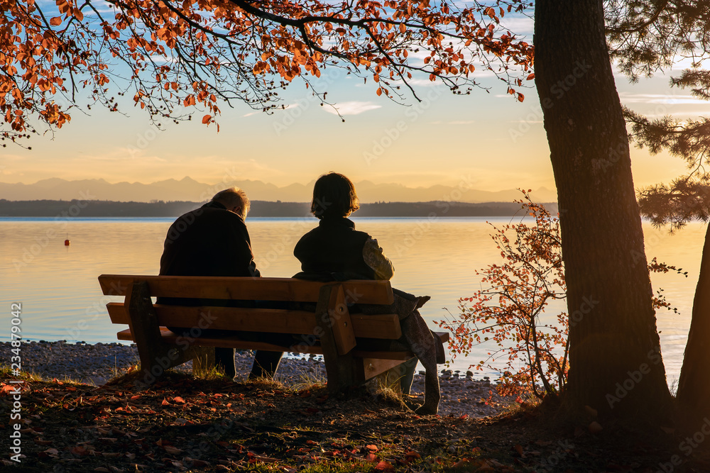 old couple are sitting on a park bench on a lake and look at the ...