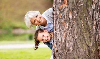 Wall Mural - family, leisure and people concept - happy grandmother and granddaughter peeking out tree at summer park