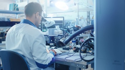 Wall Mural - Electronics Factory Worker in White Work Coat Inspects a Printed Circuit Board Through a Digital Microscope. High Tech Factory Facility.