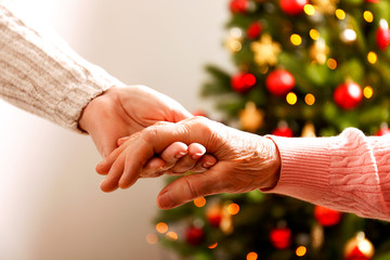 Elderly woman celebrating Christmas at home, with decorated holiday pine tree on background. Old lady at nursing home. Close up, copy space, cropped shot.