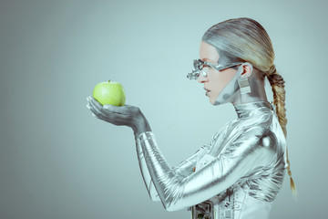 side view of cyborg holding and examining green apple isolated on grey, future technology concept